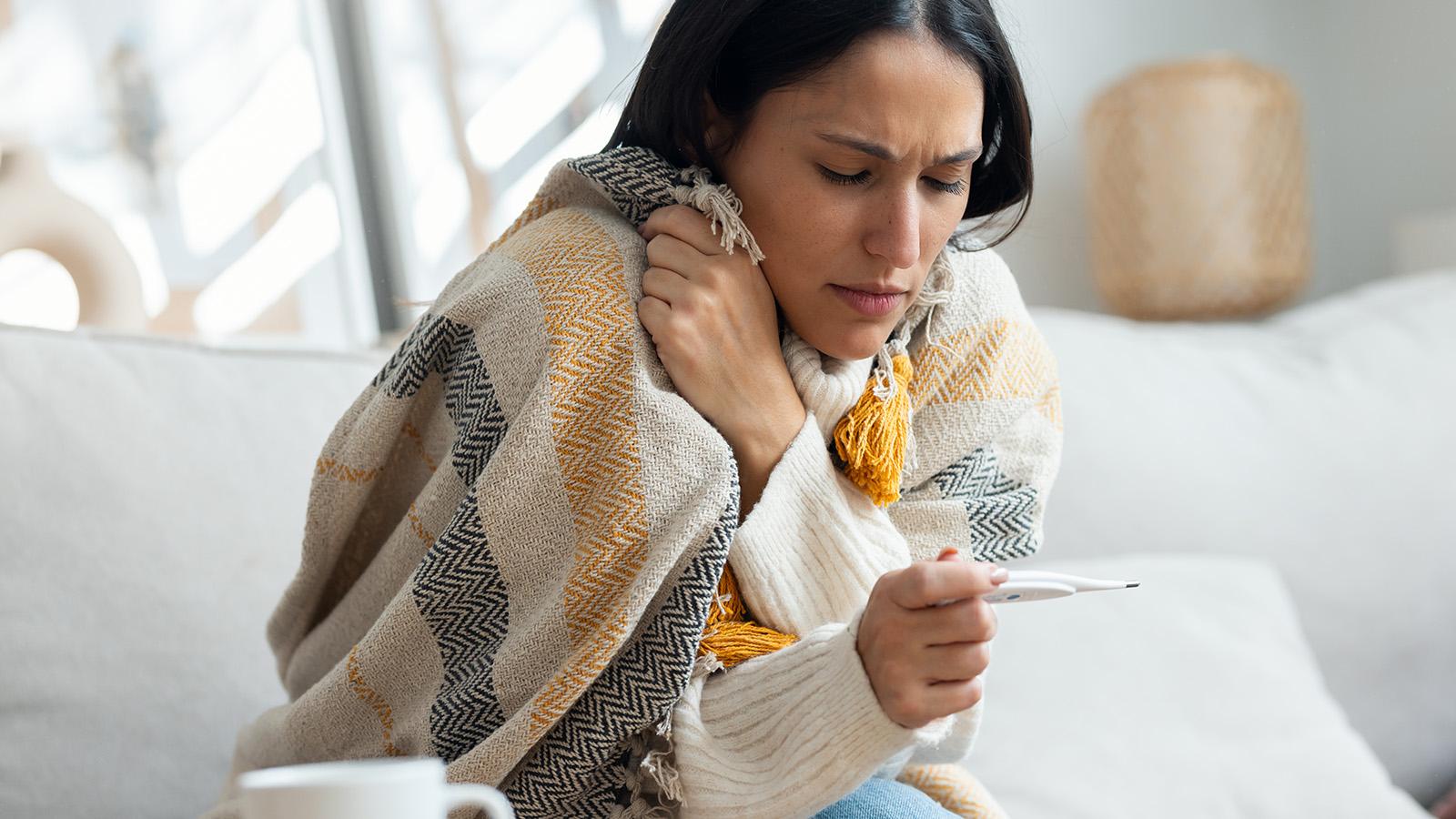 Frau liegt mit Decke auf dem Sofa und misst ihre Körpertemperatur
