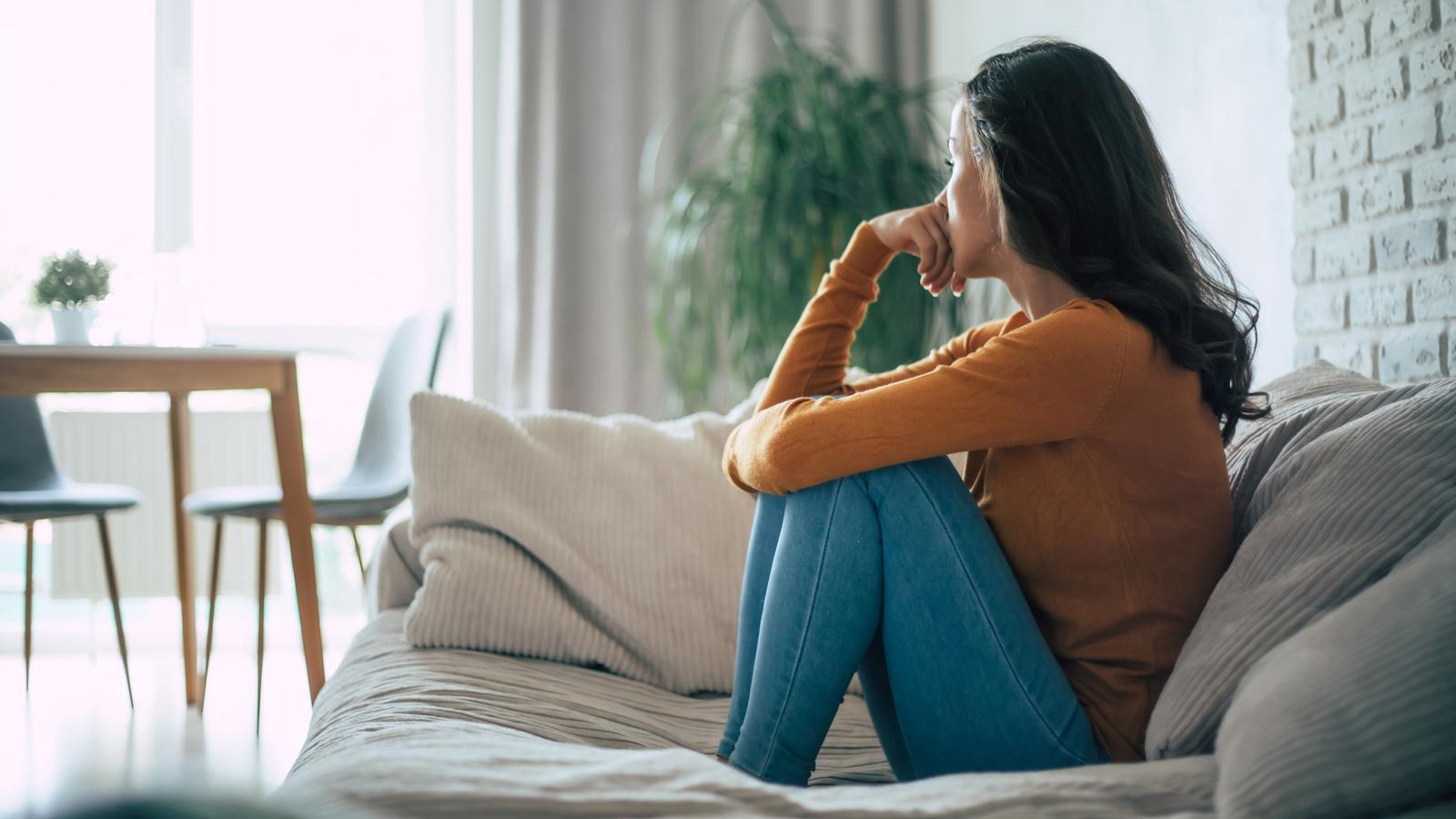 Frau sitzt nachdenklich auf der Couch und blickt Richtung Fenster