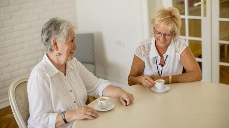 Ältere Dame sitzt mit ihrer Pflegerin zu Hause am Tisch und trinkt Kaffee