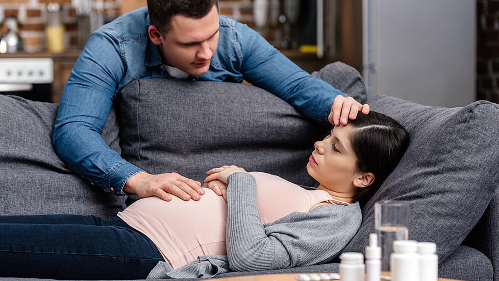 Schwangere Frau liegt am Sofa, vor ihr stehen Medikamente und ihr Mann kümmert sich um sie. 