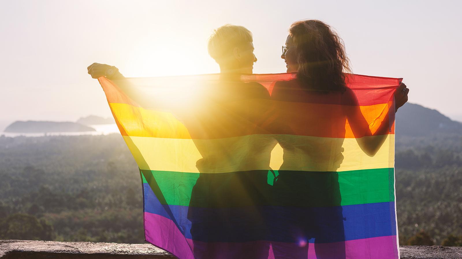 Junges lesbisches Pärchen mit Regenbogenfahne, Sonnenschein im Hintergrund