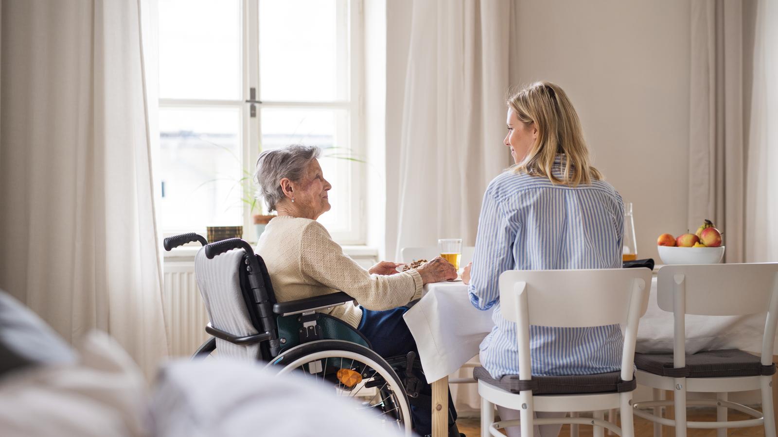 Eine ältere Frau im Rollstuhl mit einem Gesundheitsbesucher, der zu Hause am Tisch sitzt.