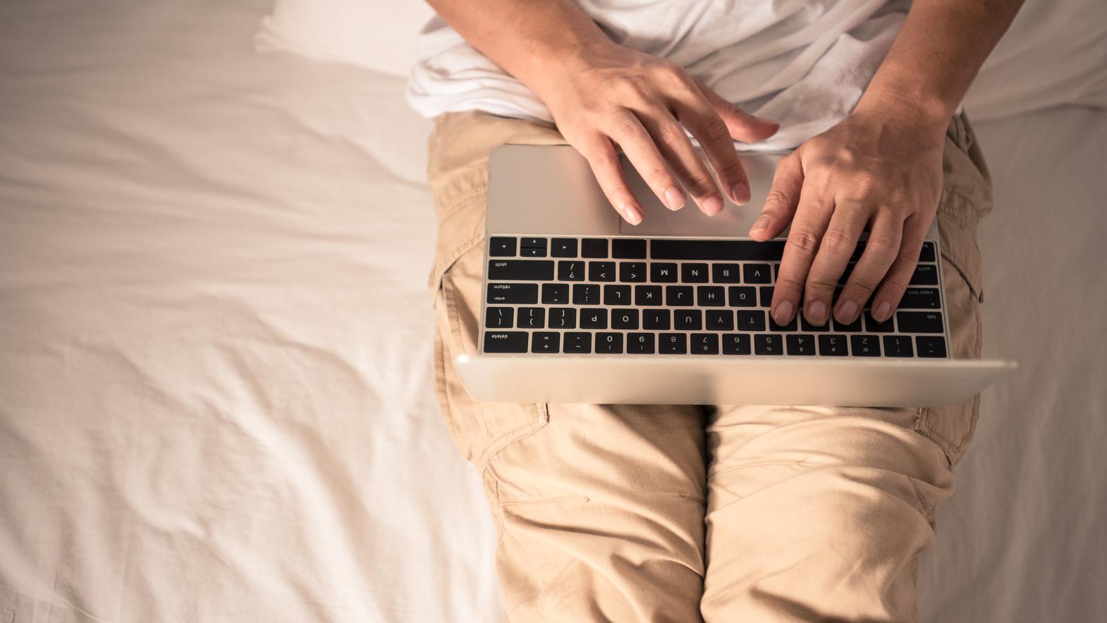 Mann sitzt mit Laptop auf dem Bett.