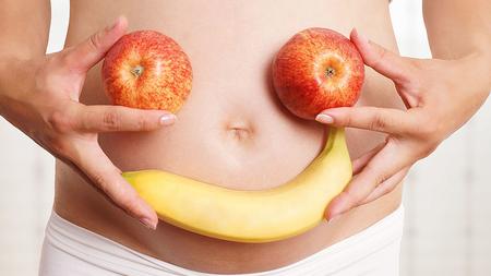 Schwangere Frau hält ein Gesicht aus Äpfeln und Banane vor ihrem Bauch