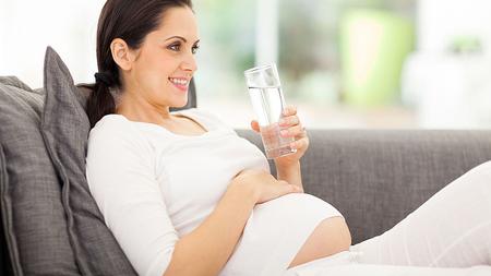 Schwangere Frau sitz auf der Couch und trinkt ein Glas Wasser. 